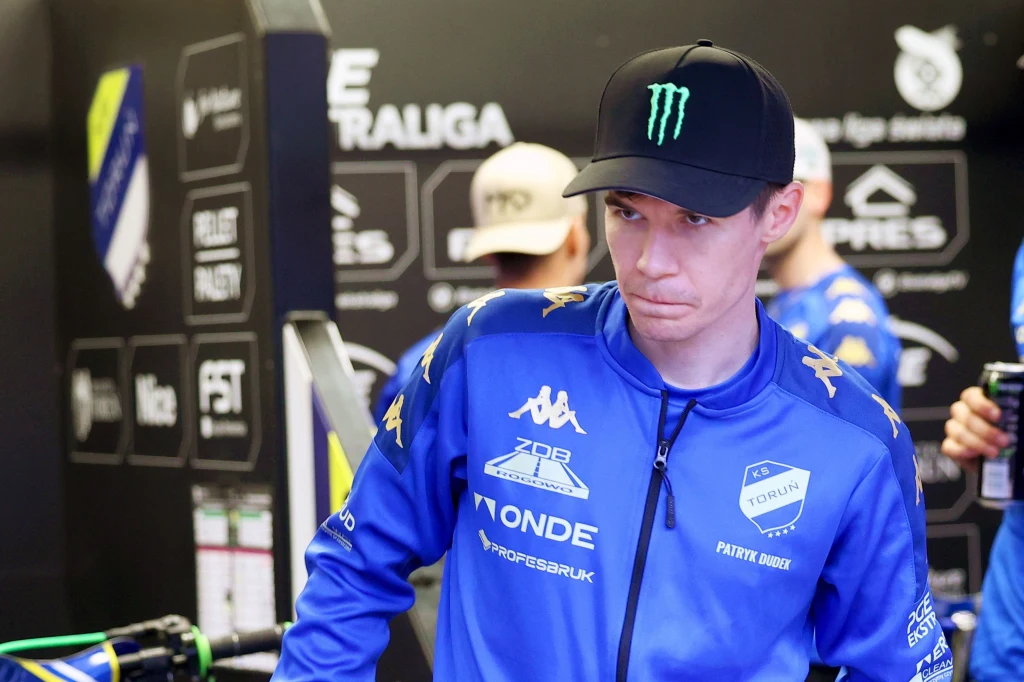 Mężczyzna w niebieskim sportowym stroju z licznymi logotypami sponsorów, na głowie czapka z daszkiem z widocznym zielonym logo, tło stanowią napisy i logo wskazujące na miejsce sportowej rywalizacji.