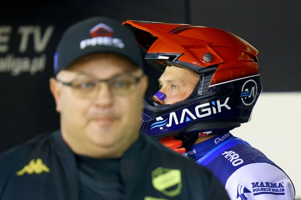 Zawodnik żużlowy w kasku i kombinezonie drużynowym stoi z boku, a na pierwszym planie znajduje się nierozpoznawalny mężczyzna w okularach i czapce, lekko rozmazany.