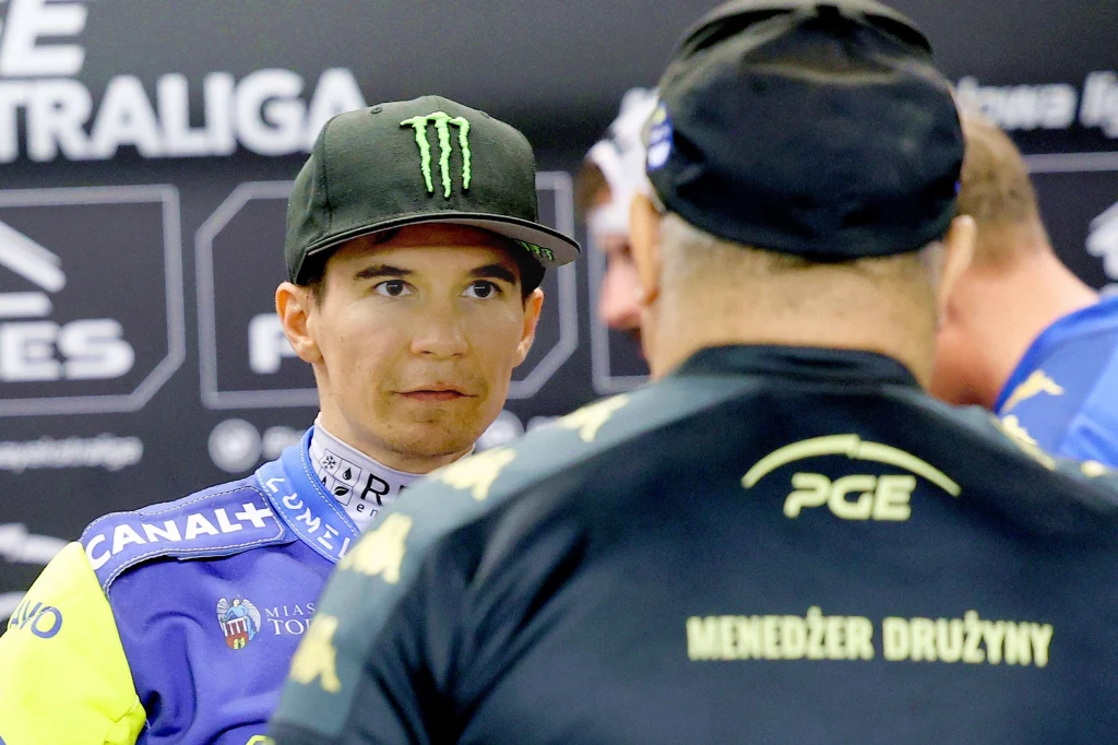 Patryk Dudek rozmawia z Piotrem Baronem Zawodnik żużlowy w pełnym stroju sportowym oraz czapce z logo sponsora stoi naprzeciwko menedżera drużyny w ubraniu z napisem PGE na plecach, tło z logo sponsorów i nazwami ligi.