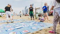 Grupa dzieci i dorosłych bawi się na plaży, uczestnicząc w grze z dużymi kartami umieszczonymi na piasku, na których znajdują się obrazki różnych przedmiotów i owoców, wokół widoczne są flagi i namioty imprezy plenerowej.