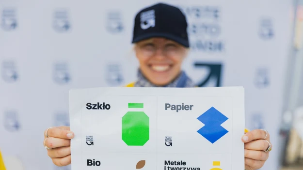 Uśmiechnięta osoba w czapce z daszkiem trzyma ulotkę edukacyjną z wyraźnymi symbolami i napisami dotyczącymi segregacji odpadów: szkło, papier, bio oraz metale i tworzywa sztuczne.