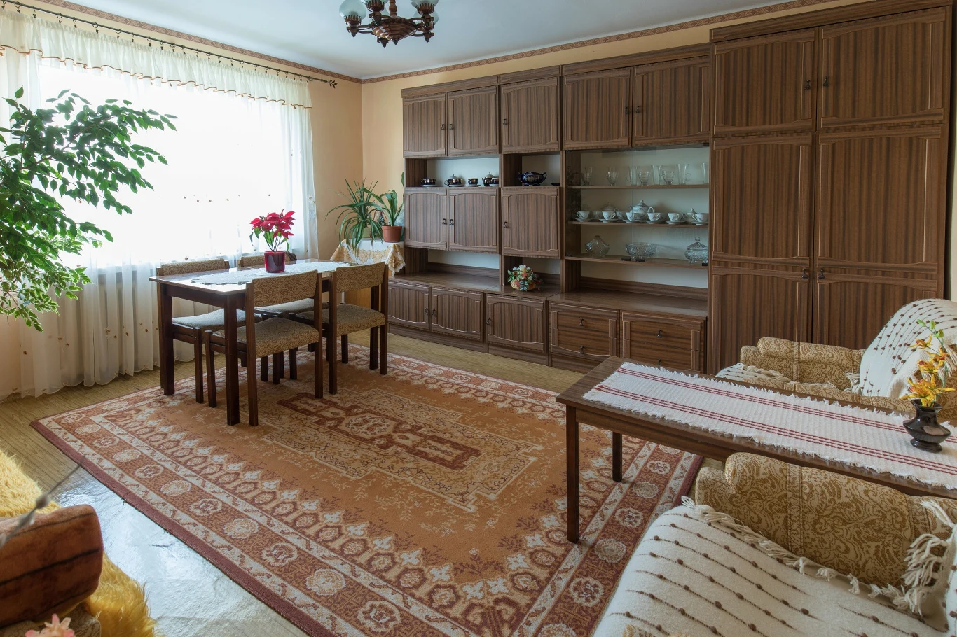 Meblościanki różniły się frontami Salon urządzony w stylu z lat 80., z segmentem meblowym przy ścianie, stołem i czterema krzesłami, dużym dywanem na podłodze oraz rozłożystą rośliną przy oknie, przez które wpada naturalne światło.