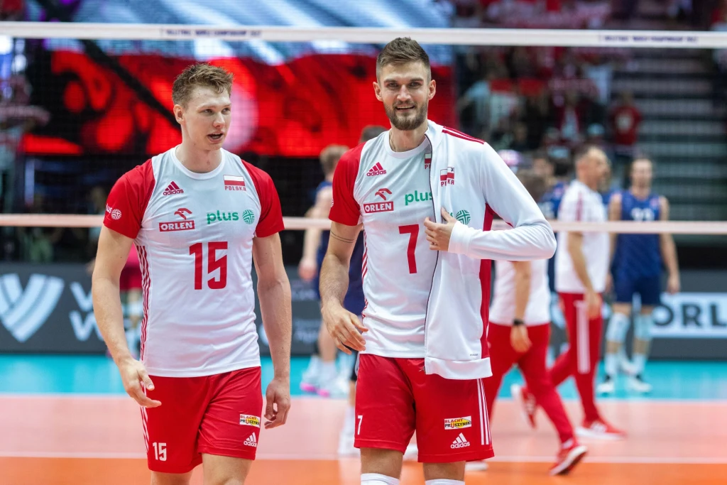 Jakub Kochanowski i Karol Kłos Dwóch siatkarzy reprezentacji Polski w czerwono-białych strojach drużynowych idzie po boisku podczas meczu siatkówki, w tle widoczni pozostali zawodnicy oraz sportowa hala.