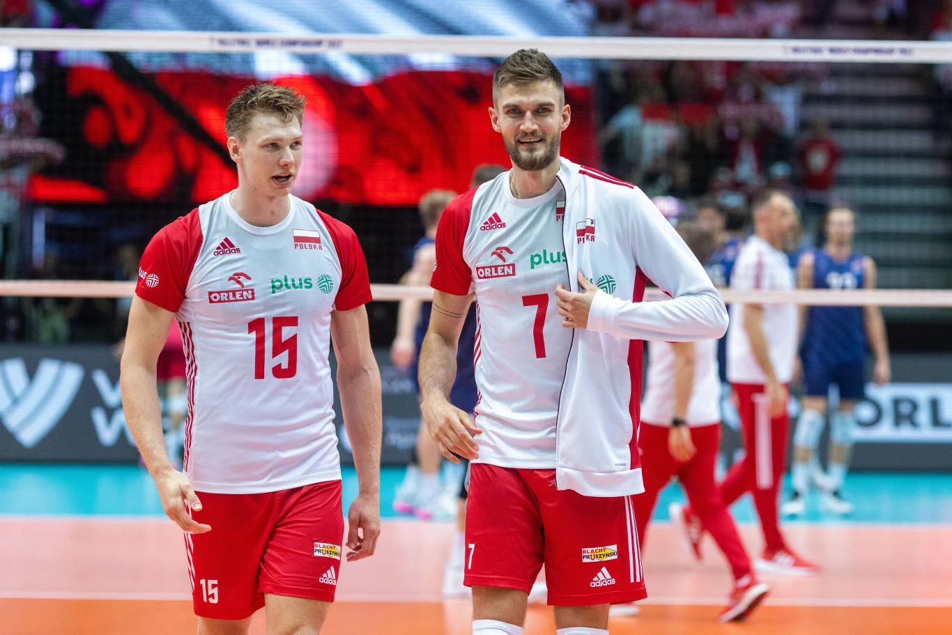 Dwóch siatkarzy reprezentacji Polski w czerwono-białych strojach drużynowych idzie po boisku podczas meczu siatkówki, w tle widoczni pozostali zawodnicy oraz sportowa hala.