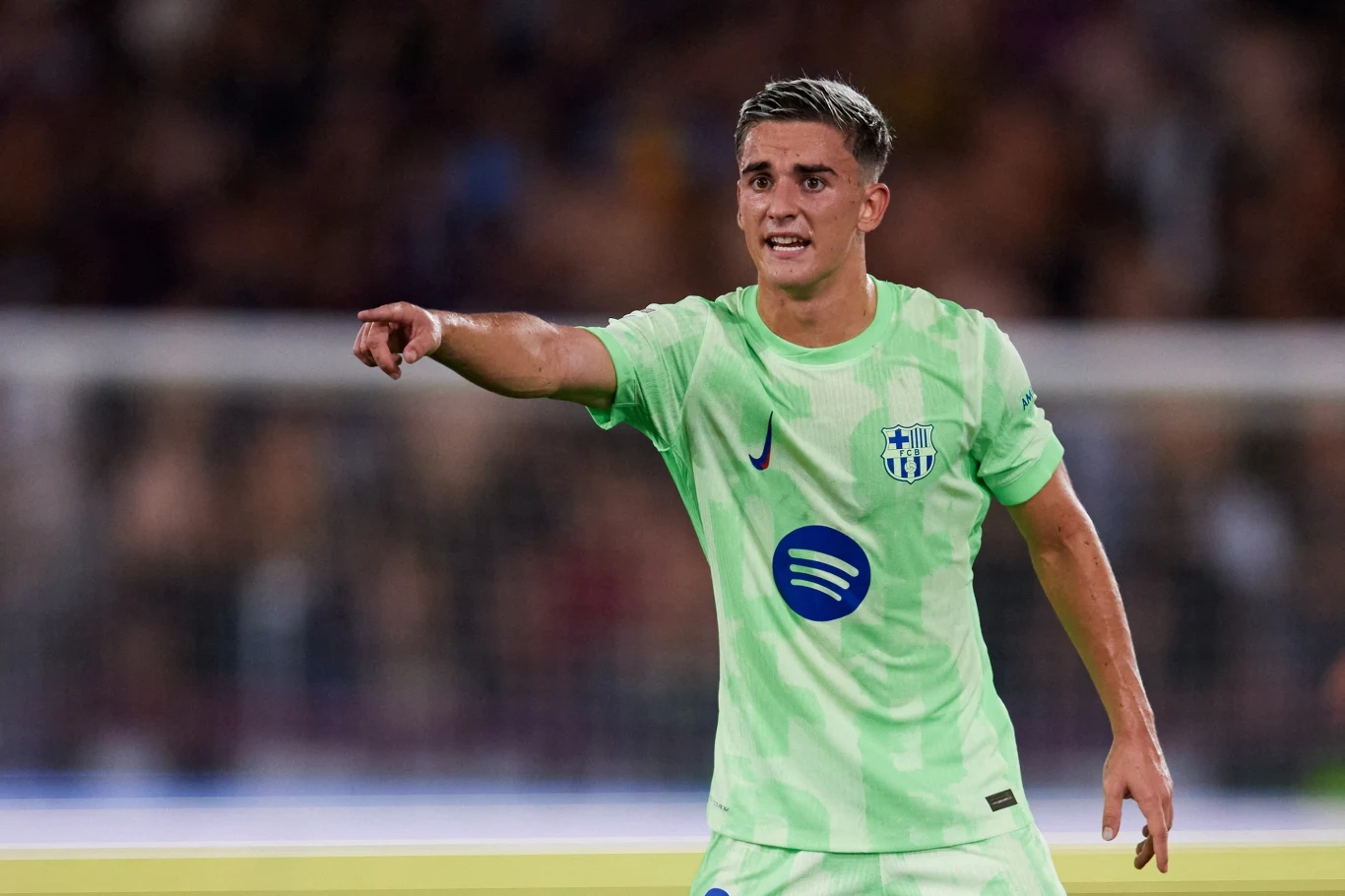 Młody piłkarz w jasnozielonej koszulce Barcelony, z krótkimi włosami, wskazuje ręką kierunek podczas meczu, tło zamazane z widownią stadionu.