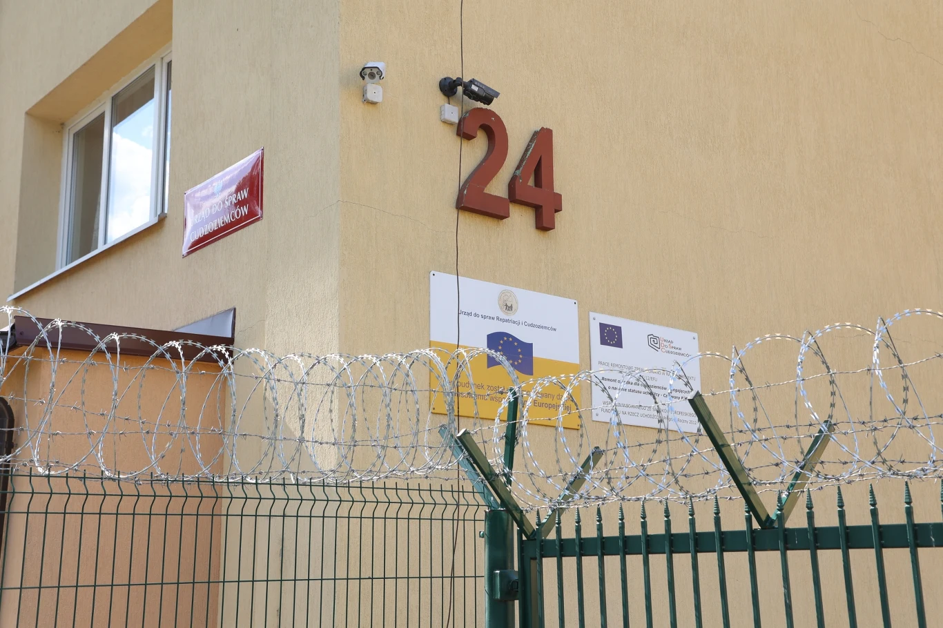 RPO ostrzega przed przepełnieniem ośrodków dla cudzoziemców Numer budynku 24 wyeksponowany na żółtej elewacji, tablice informacyjne z logotypami Unii Europejskiej i innych instytucji, wysoki zielony płot zakończony drutem kolczastym, kamera monitoringu oraz okno.