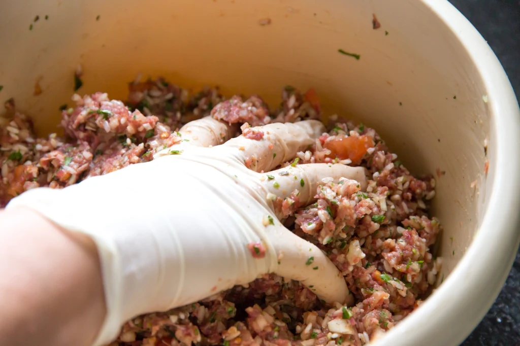 Ręka w lateksowej rękawiczce zagniata w misce mieszankę mielonego mięsa z posiekanymi warzywami i przyprawami, składniki dobrze połączone i gotowe do dalszej obróbki kulinarnej.