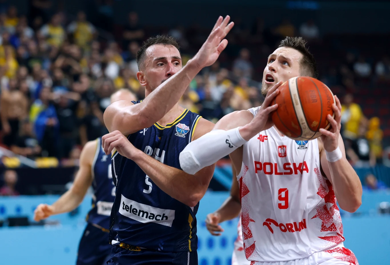 Mateusz Ponitka (P) w meczu z Bośnią i Hercegowiną Dwóch koszykarzy w akcji podkoszowej podczas meczu, zawodnik w białej koszulce z napisem Polska trzyma piłkę i przygotowuje się do rzutu, natomiast zawodnik w granatowej koszulce drużyny przeciwnej próbuje go zablokować; w tle rozmazana publiczność i n...