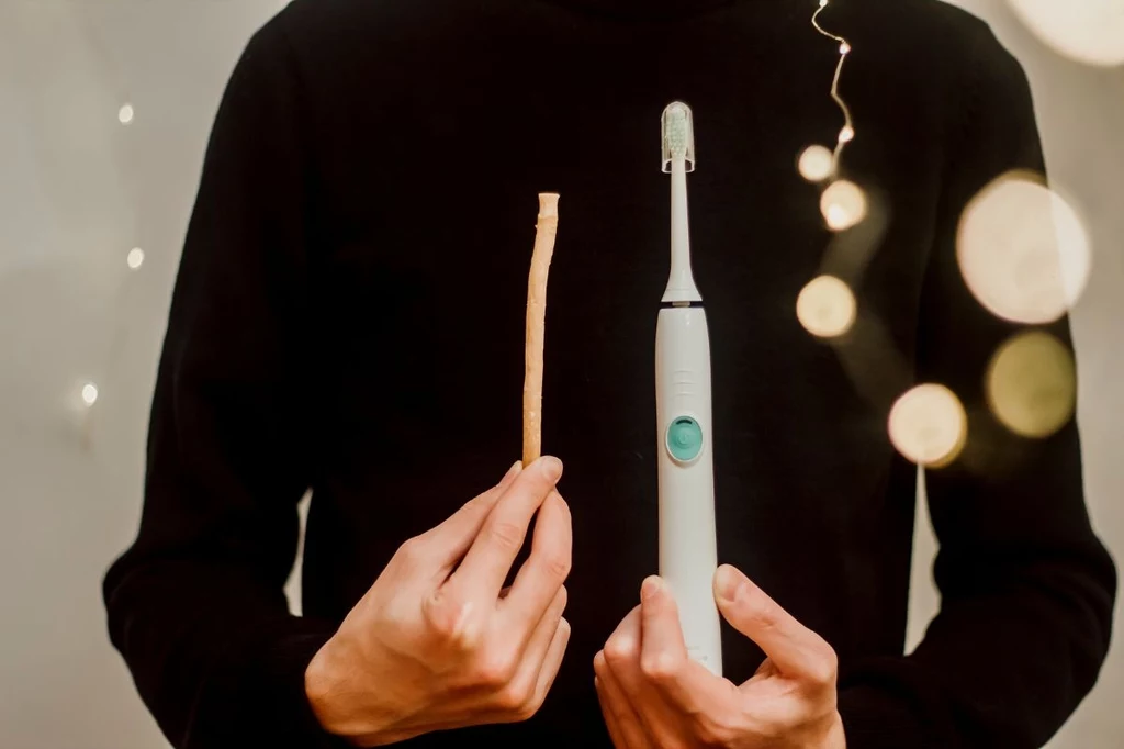 Miswak w porównaniu do szczoteczki elektrycznej wygląda nieco prymitywnie Porównanie tradycyjnej gałązki do czyszczenia zębów oraz nowoczesnej szczoteczki elektrycznej trzymanych przez osobę ubraną na czarno.