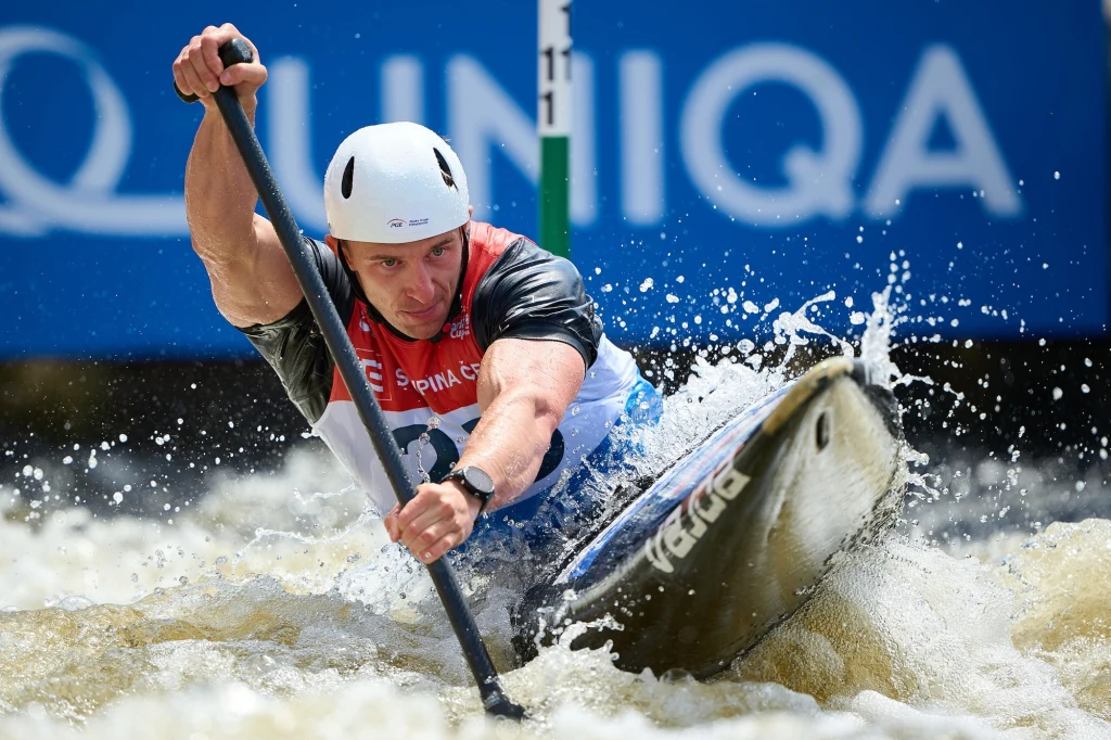 Zawodnik w kasku i kamizelce startowej intensywnie wiosłuje kajakiem, pokonując bystrą wodę na torze slalomowym, wokół rozpryskują się fale.