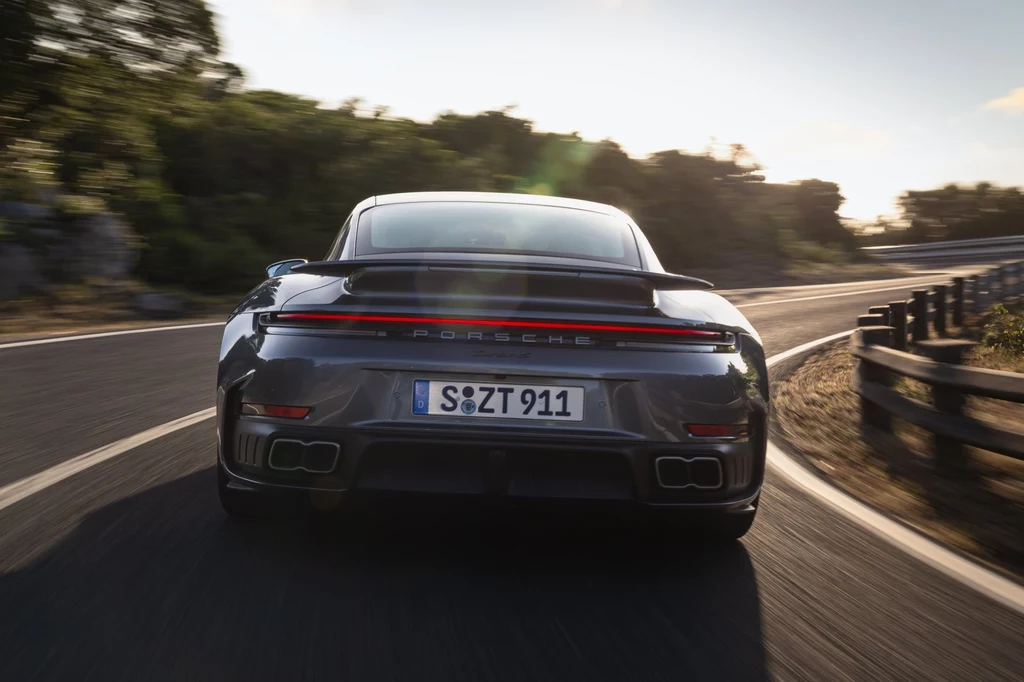 Współczynnik oporu powietrza w nowym Porsche 911 Turbo S został poprawiony o 10 proc. względem poprzednika. Srebrny samochód sportowy Porsche 911 jedzie pustą, krętą drogą otoczoną zielenią i barierkami ochronnymi, światło słoneczne subtelnie odbija się od karoserii.