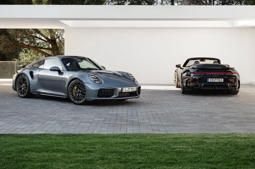 Porsche 911 Turbo S dostępne jest jako coupe i kabriolet. Ceny startują od 1 373 000 zł za pierwszą wersję i od 1 442 000 zł za drugą. Dwa sportowe samochody zaparkowane na podjeździe przed nowoczesnym białym budynkiem, w tle zieleń drzew
