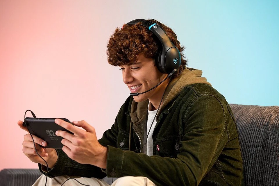 Turtle Beach Airlite Fit Młody mężczyzna z kręconymi włosami siedzi na kanapie, gra na konsoli przenośnej i nosi słuchawki z mikrofonem, wygląda na skoncentrowanego i zadowolonego.