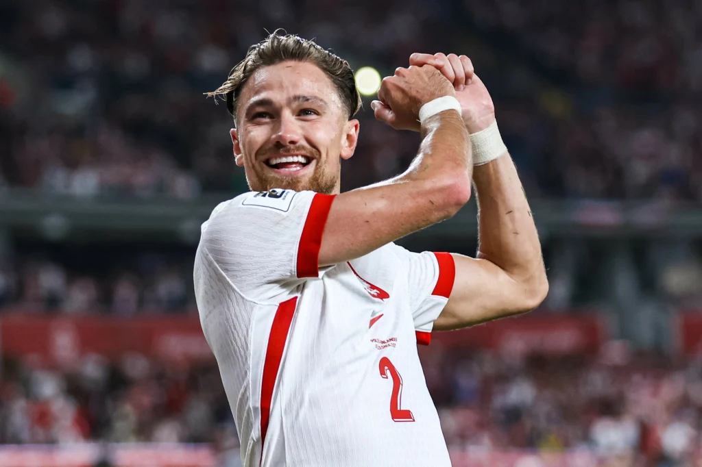 Piłkarz w białym stroju z czerwonymi elementami, uśmiechnięty, klaska w dłonie, w tle rozmyty stadion i kibice.