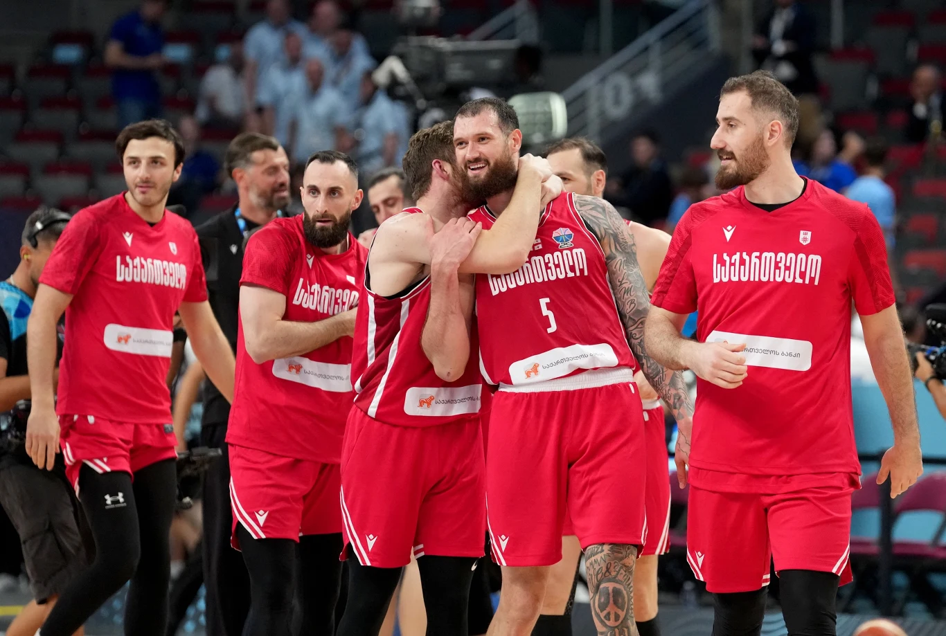 Grupa koszykarzy w czerwonych strojach cieszy się po zakończonym meczu na hali sportowej, kilku zawodników obejmuje się, na twarzach widać radość i dumę, w tle widoczni są kibice i trybuny.