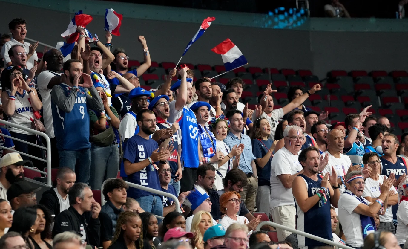 Tłum kibiców ubranych w niebieskie i białe koszulki z napisem 'France', ludzie machają francuskimi flagami, dopingują i wyrażają entuzjazm podczas wydarzenia sportowego w hali.
