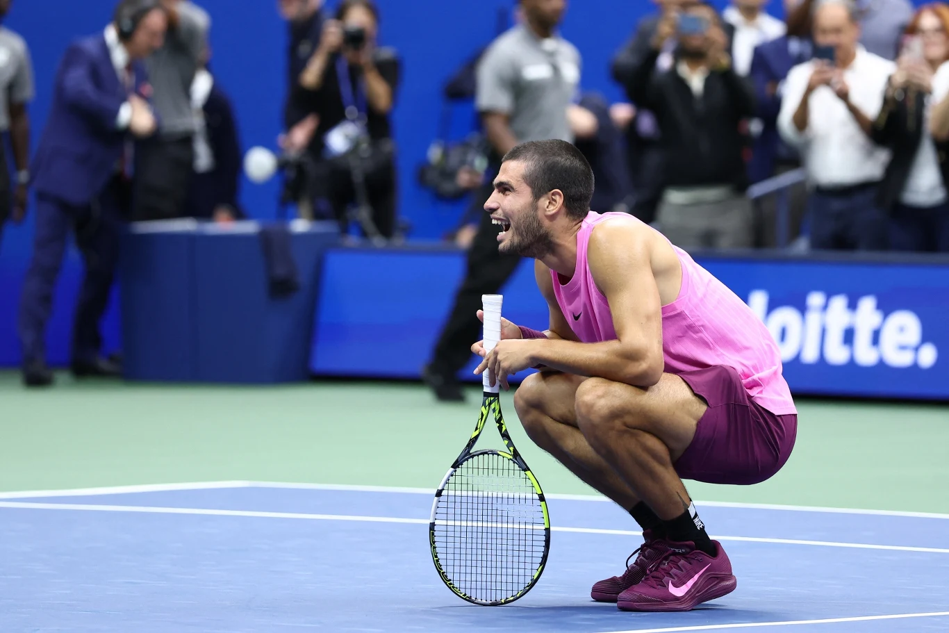 Tenisista ubrany w różowy strój kuca na korcie, trzymając rakietę tenisową i uśmiechając się szeroko, w tle rozmazani kibice oraz fotografowie robiący zdjęcia.