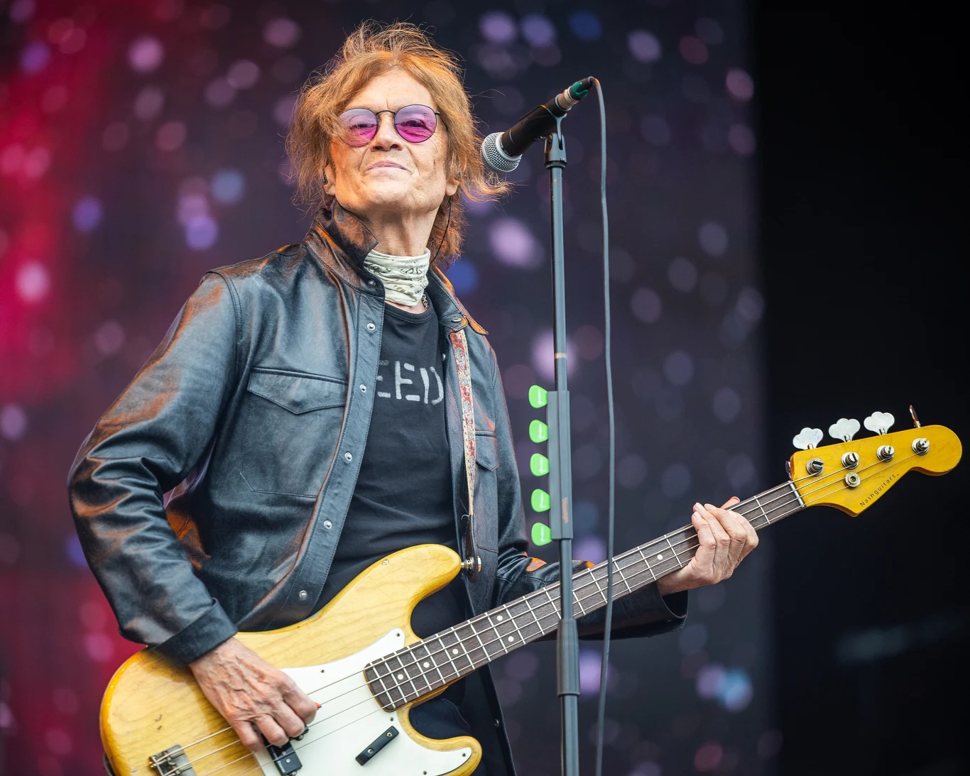 Glenn Hughes przygotował nowy materiał Mężczyzna w okularach przeciwsłonecznych grający na gitarze basowej, ubrany w czarną skórzaną kurtkę i ciemny t-shirt, stoi na scenie z mikrofonem i kolorowym, rozmytym tłem.