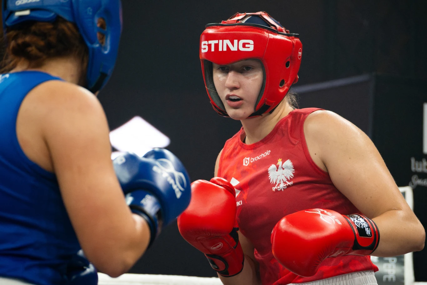 Dwie kobiety w kaskach ochronnych boksujące się na ringu, jedna ubrana w czerwony strój z godłem Polski, skoncentrowana na przeciwniczce w niebieskim stroju.