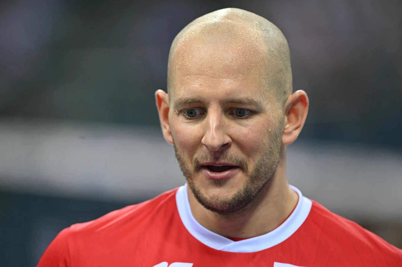 Łysy mężczyzna w czerwonej koszulce sportowej z białym kołnierzykiem stoi w lekkim uśmiechu, w tle widoczne rozmycie.