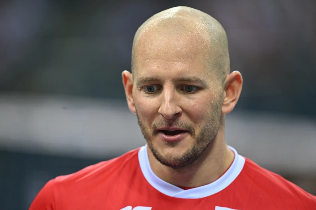 Łysy mężczyzna w czerwonej koszulce sportowej z białym kołnierzykiem stoi w lekkim uśmiechu, w tle widoczne rozmycie.