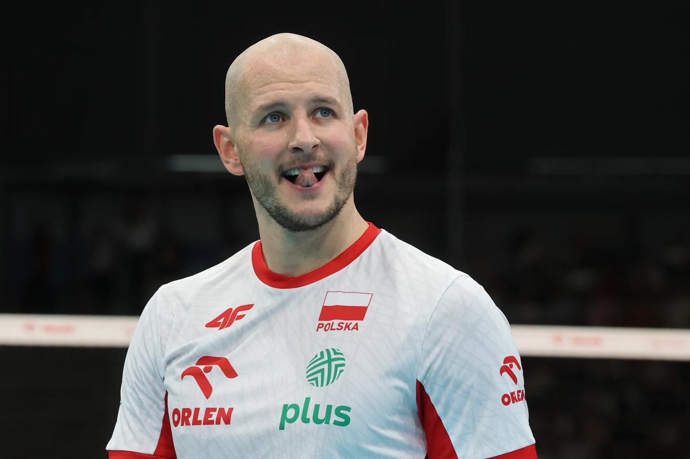 Siatkarz w koszulce reprezentacji Polski z logo sponsorów, uśmiechnięty i z wystawionym lekko językiem, stoi na tle hali sportowej.