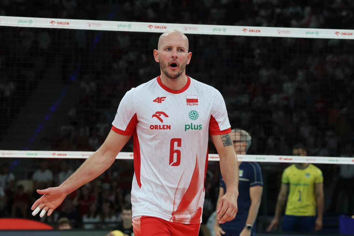 Polski siatkarz w białoczerwonym stroju z numerem 6 na piersi gestykuluje na boisku podczas meczu, w tle siatka oraz zawodnicy drużyny przeciwnej w żółtozielonych strojach.