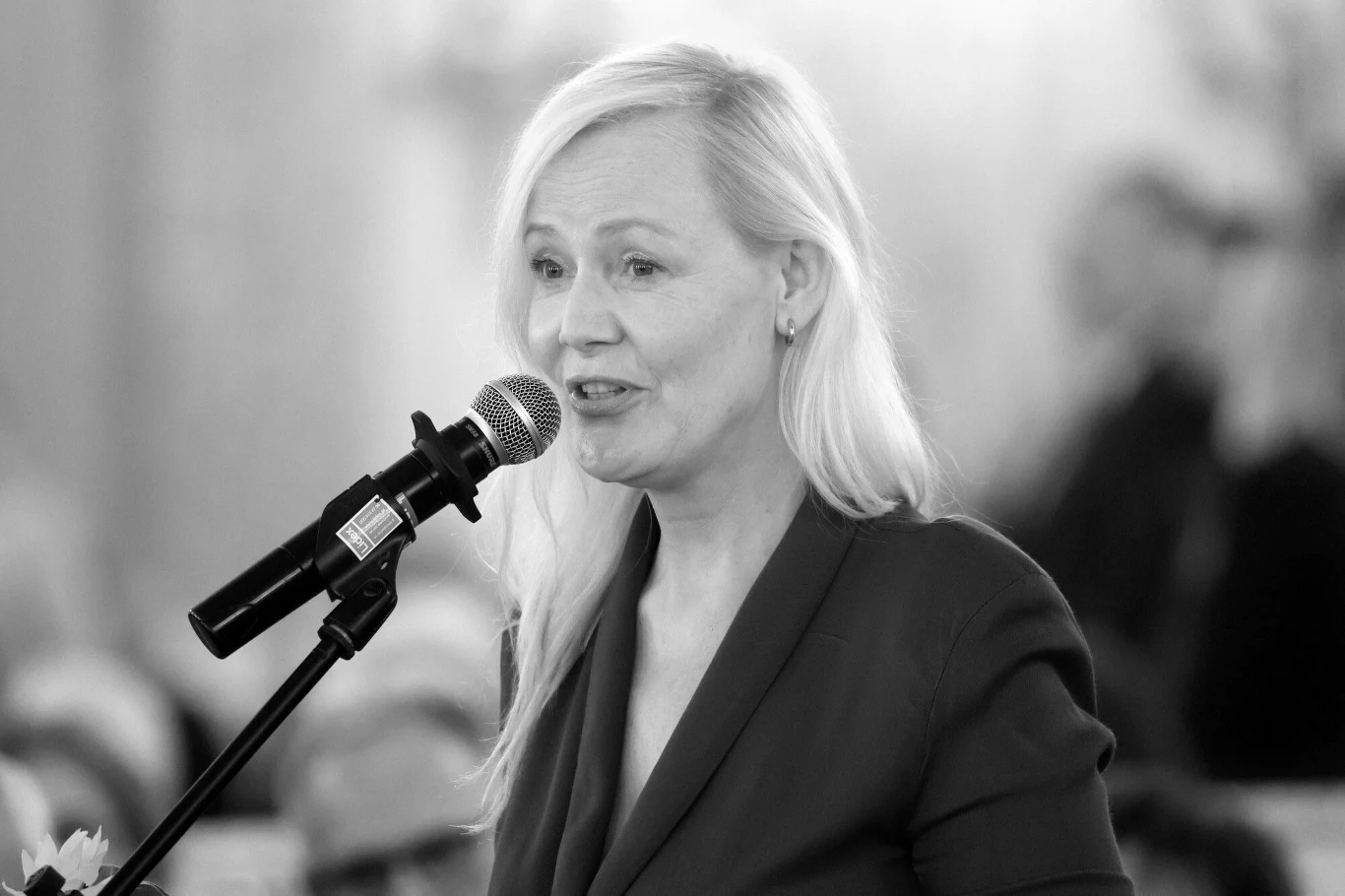 Katarzyna Stoparczyk zginęła w wypadku samochodzowym Kobieta z jasnymi włosami przemawia do mikrofonu w eleganckim stroju, skupiona na swojej wypowiedzi, w tle rozmyte sylwetki osób.