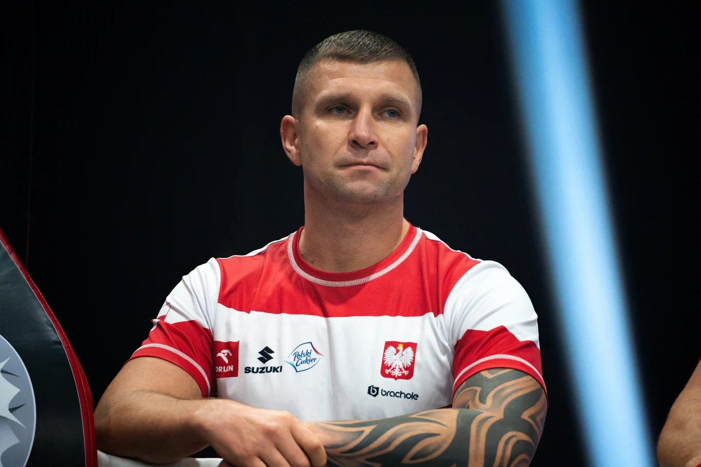 Tomasz Dylak, trener reprezentacji Polski Mężczyzna w sportowej koszulce reprezentacji Polski z widocznymi tatuażami na ramionach siedzi przy stoliku, patrząc z powagą przed siebie.