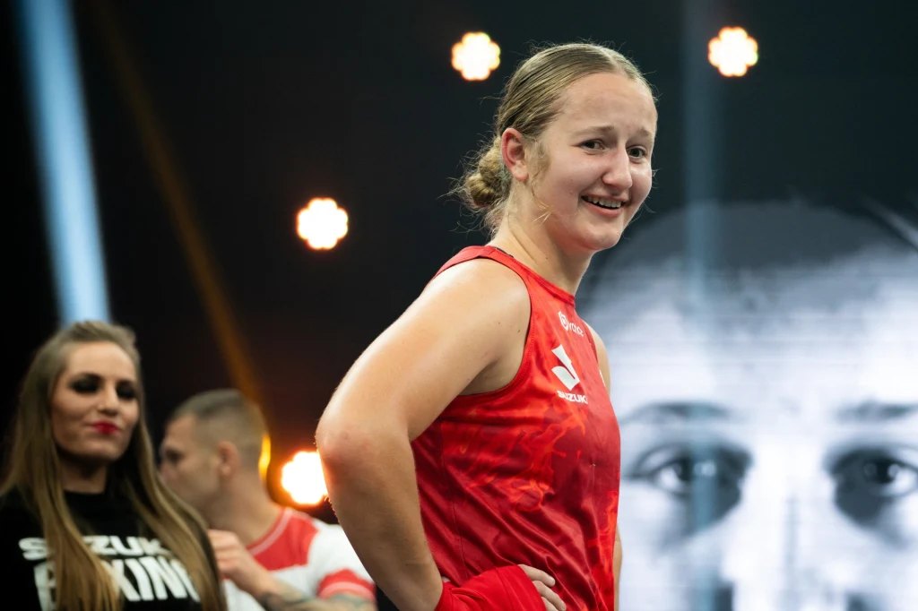 Emilia Koterska Kobieta ubrana w czerwony strój bokserski stoi na ringu, lekko uśmiechnięta, w tle osoby towarzyszące i rozmyte światła.