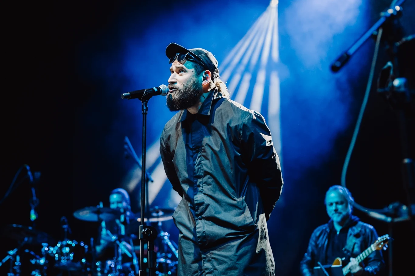 Fama 2025: Sławek Przybył podczas koncertu "Tu był Grzegorz z Ciechowa" Mężczyzna z brodą i czapką śpiewa do mikrofonu podczas koncertu na scenie, za nim zespół muzyczny gra na instrumentach, a całość oświetlona jest niebieskimi reflektorami.