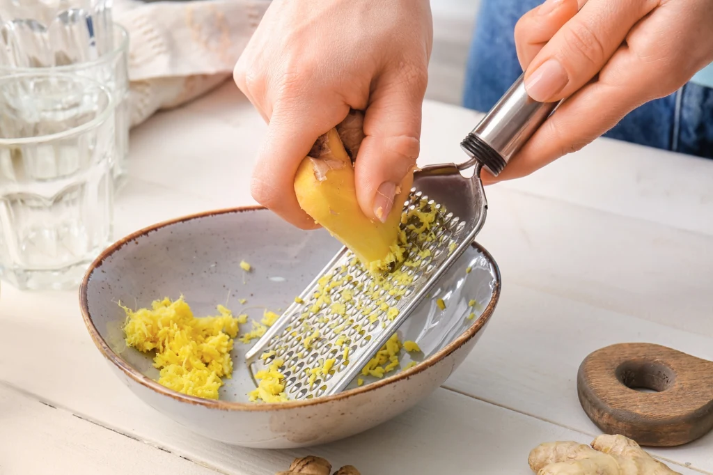 Tarte świeże kawałki imbiru na metalowej tarce trzymanej nad ceramiczną miską, obok na stole leżą plastry imbiru oraz szklanki w tle