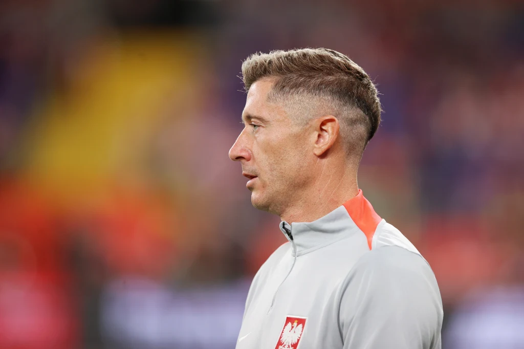 Profil mężczyzny w sportowej kurtce z godłem Polski, rozmyte wielokolorowe tło stadionu.