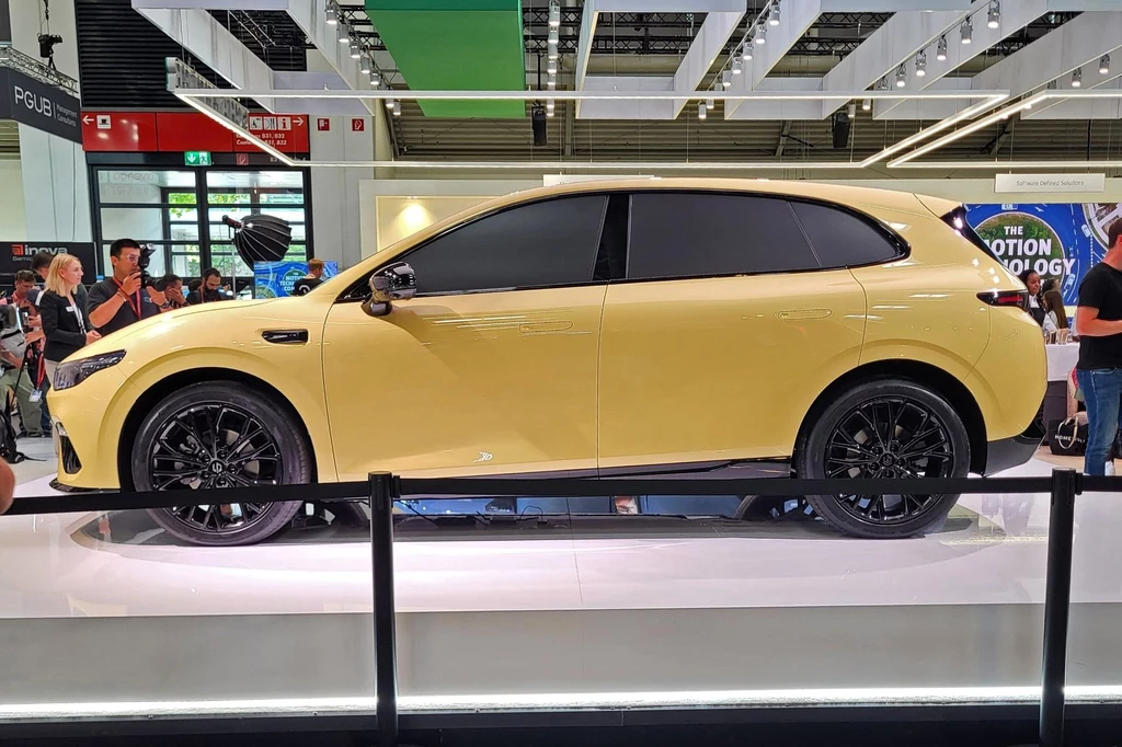 Jasnożółty nowoczesny SUV z czarnymi detalami, stojący na ekspozycji w przestronnej hali targowej, otoczony przez odwiedzających i osadzony w jasnym, nowoczesnym wnętrzu.