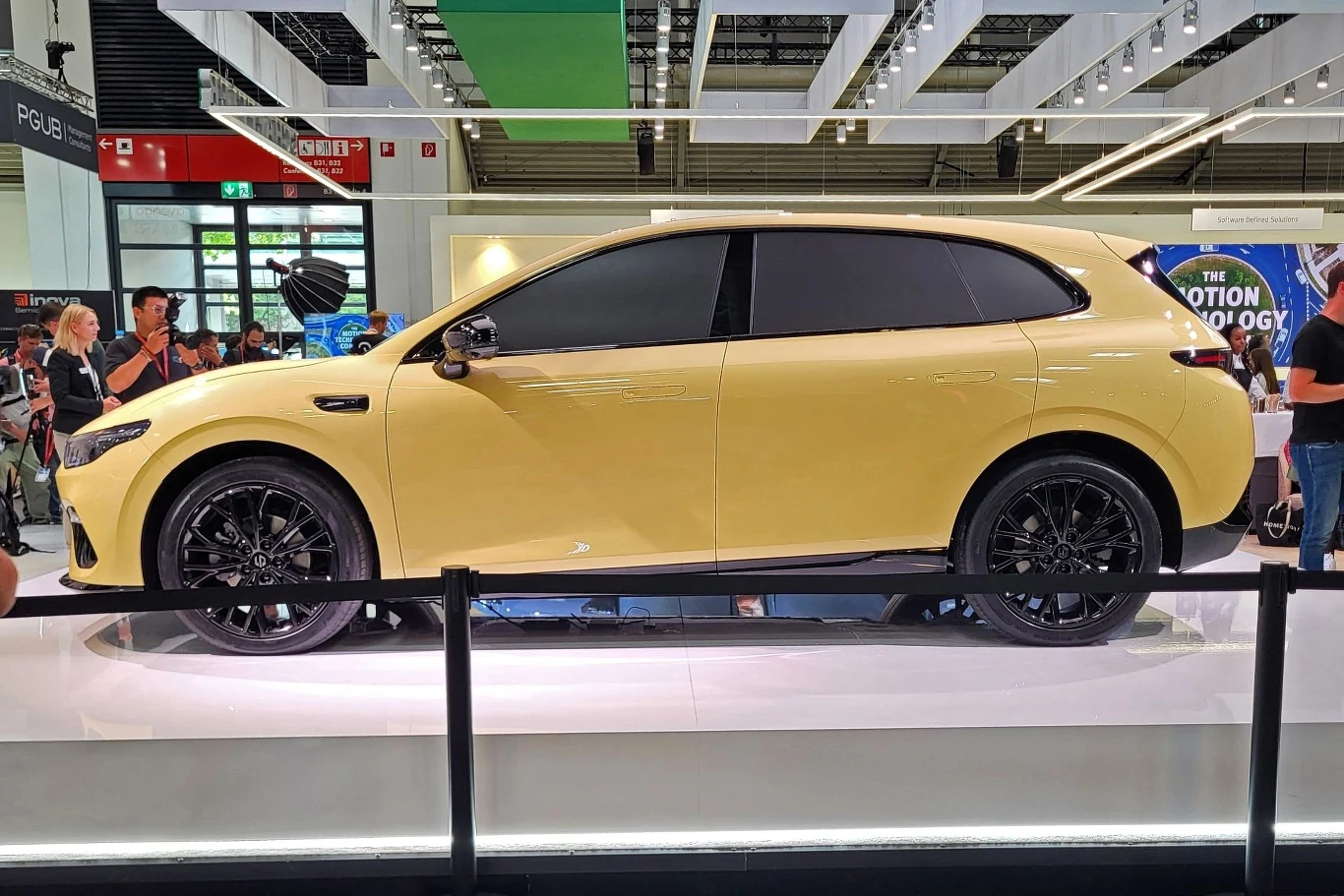 Jasnożółty nowoczesny SUV z czarnymi detalami, stojący na ekspozycji w przestronnej hali targowej, otoczony przez odwiedzających i osadzony w jasnym, nowoczesnym wnętrzu.