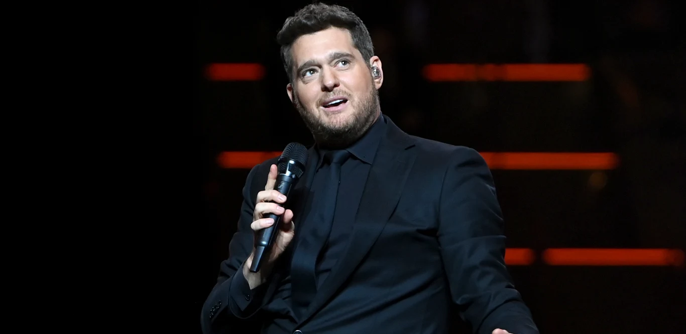 Michael Bublé kończy 50 lat Mężczyzna ubrany w ciemny garnitur trzyma mikrofon i śpiewa na scenie, za nim ciemne tło z kilkoma poziomymi liniami świetlnymi.