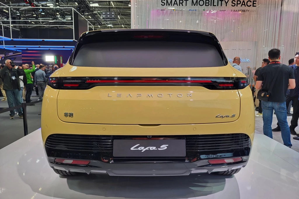 Tył żółtego samochodu elektrycznego marki Leapmotor model Capa 5 ustawiony na białej platformie wystawowej, wokół pojazdu stoją odwiedzający targi motoryzacyjne, a w tle widoczne hasła promujące nowoczesną mobilność.