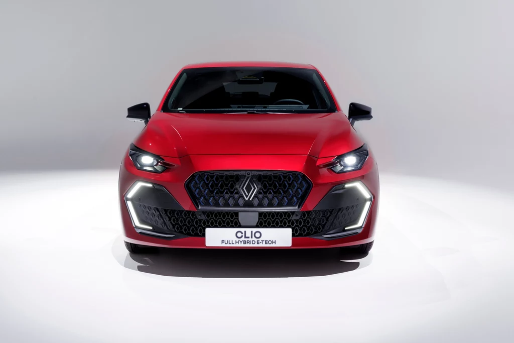 Nowe spojrzenie przodu Clio zawdzięcza między innymi odmienionym, węższym reflektorom, dzięki którym wygląda bardziej zadziornie Czerwony samochód osobowy Renault Clio prezentowany od frontu na jasnym, neutralnym tle, widoczne nowoczesne reflektory, wyrazisty grill oraz tablica z napisem Clio full hybrid tech.