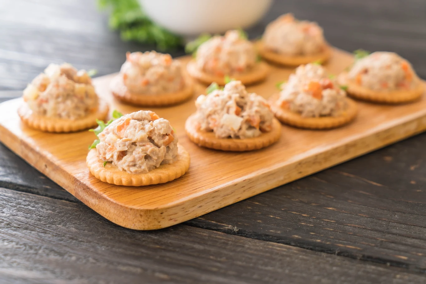 Imprezowy fingerfood - pasta rybna na krakersach Okrągłe krakersy z sałatką tuńczykową ułożone na drewnianej desce, w tle delikatnie rozmyte zielone liście.