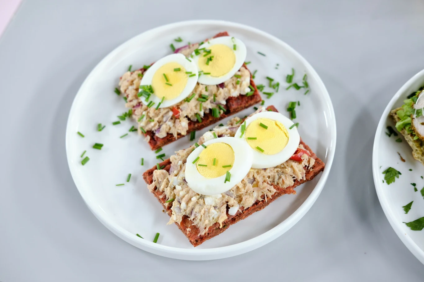 Dwie kanapki z chleba razowego posmarowane pastą z tuńczyka, udekorowane plasterkami ugotowanego na twardo jajka i świeżo posiekanym szczypiorkiem, podane na białym talerzu.