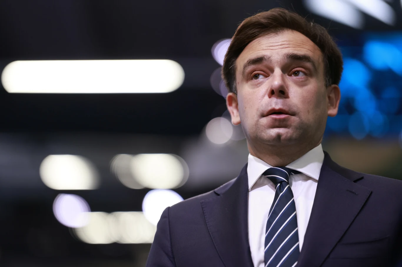 Obniżenie perspektywy ratingu Polski przez Agencję Fitch to żółta kartka dla rządzących, w tym szefa MF Andrzeja Domańskiego Mężczyzna w garniturze i krawacie patrzy w bok, tło rozmyte z jasnymi światłami.