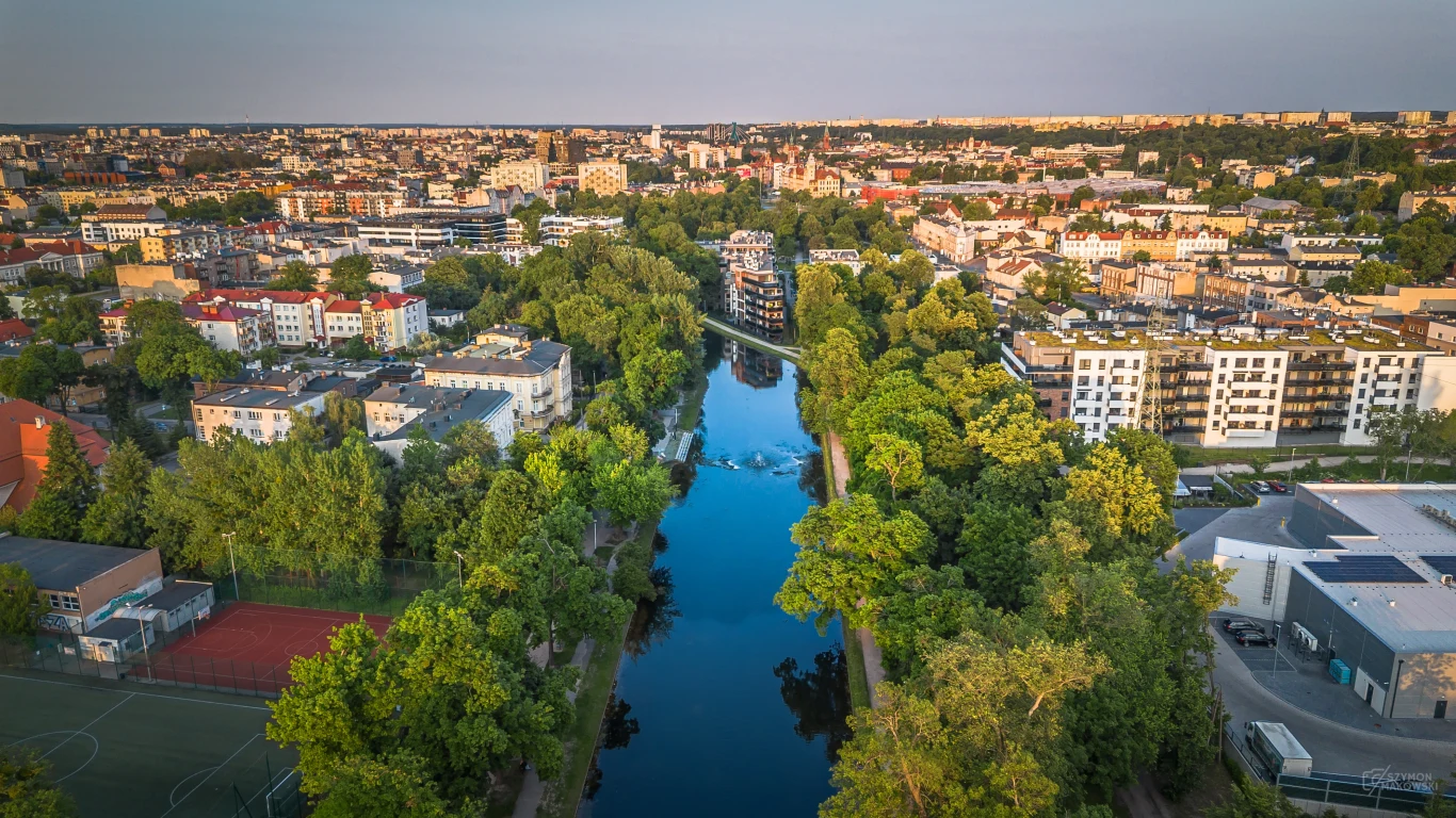 Park nad kanałem w Bydgoszczy - Fot. Miasto Bydgoszcz Panorama miasta z lotu ptaka, widoczna szeroka rzeka otoczona gęstą zielenią, po obu stronach liczne budynki mieszkalne i usługowe, w tle rozległa zabudowa wielkomiejska.