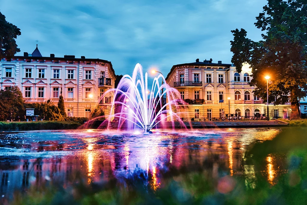 Kolorowo podświetlona fontanna pośrodku miejskiego placu otoczona klasycystycznymi kamienicami, odbicia świateł na wodzie i wieczorne niebo tworzą malowniczą scenerię.