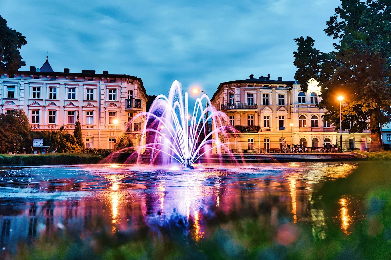 Park nad Kanałem w Bydgoszczy - Fot. Miasto Bydgoszcz Kolorowo podświetlona fontanna pośrodku miejskiego placu otoczona klasycystycznymi kamienicami, odbicia świateł na wodzie i wieczorne niebo tworzą malowniczą scenerię.