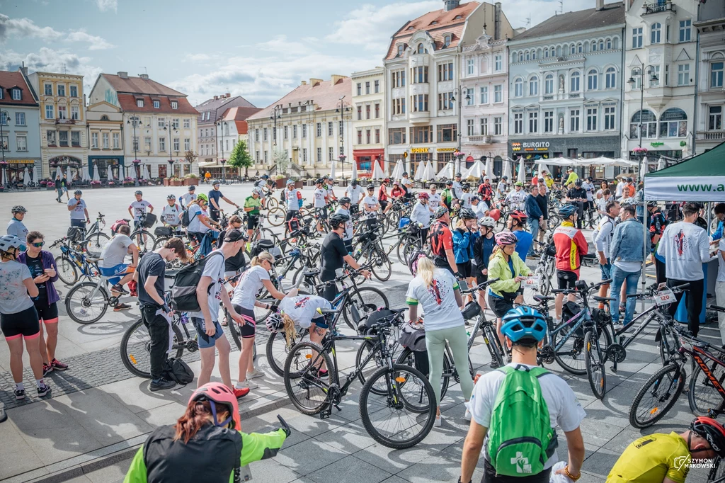 Tłum ludzi zgromadzonych na rowerach na dużym miejskim rynku w otoczeniu zabytkowych kamienic, uczestnicy ubrani sportowo, większość ma kaski i numerki na koszulkach, atmosfera wydarzenia masowego.