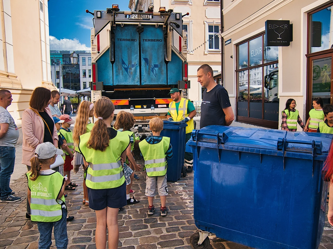 Edukacja segregacji odpadów - Fot. Miasto Bydgoszcz Grupa dzieci w odblaskowych kamizelkach wraz z opiekunami obserwuje pracowników służb komunalnych przy śmieciarce na miejskiej ulicy, między dużymi niebieskimi pojemnikami na odpady.