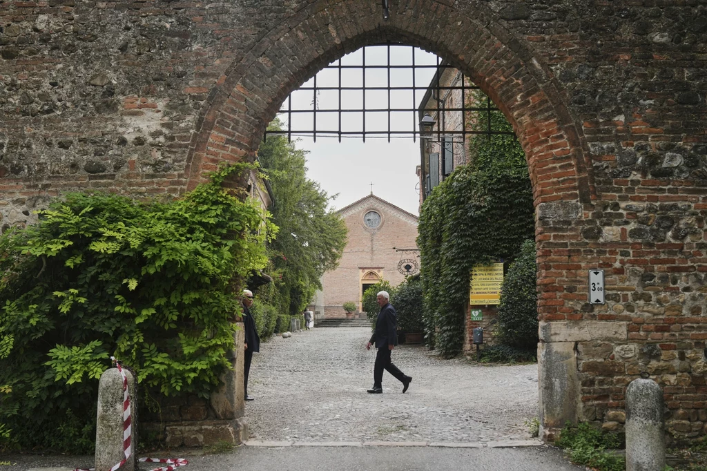 Kościół San Martino przed prywatnym pogrzebem Giorgio Armaniego Kamienna brama z kratą prowadzi na dziedziniec z brukowaną ścieżką, po której idzie mężczyzna w garniturze, w tle widoczny budynek przypominający kościół lub klasztor, otoczony zielenią i porośnięty bluszczem.