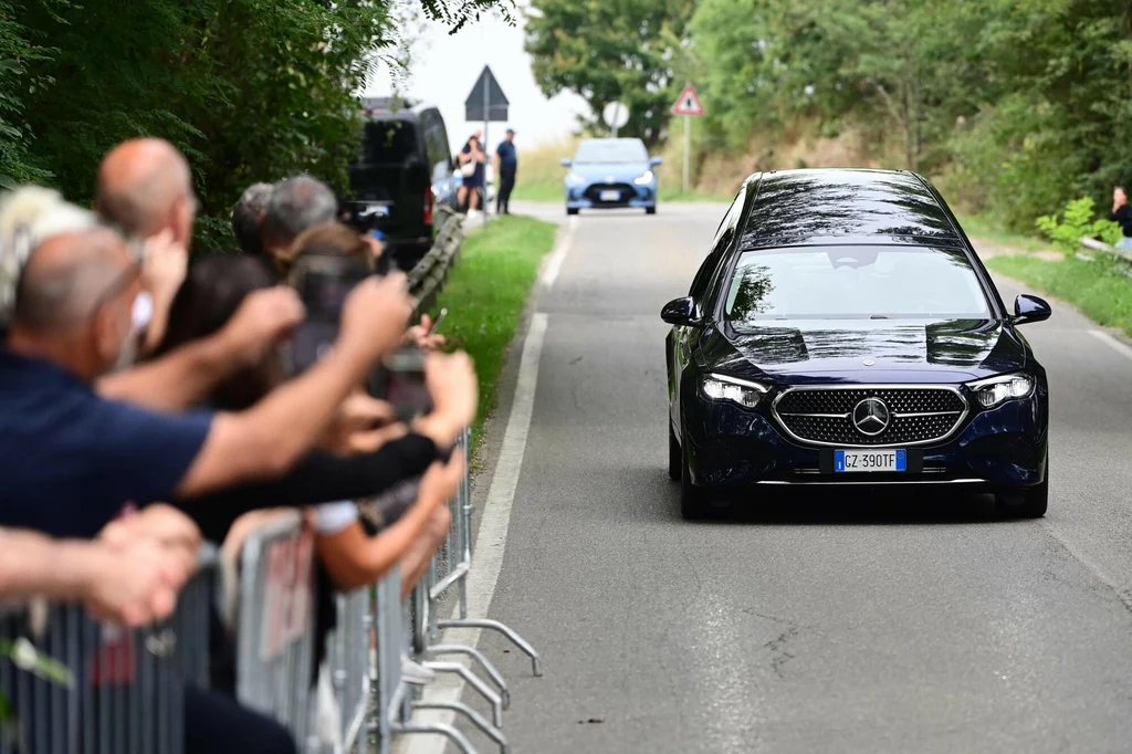 Karawan z ciałem zmarłego włoskiego projektanta mody Giorgio Armaniego Karawan marki Mercedes-Benz jedzie wąską, wiejską drogą, po lewej stronie widoczny tłum ludzi stojących za barierkami, którzy obserwują lub żegnają pojazd, w tle widoczny radiowóz.