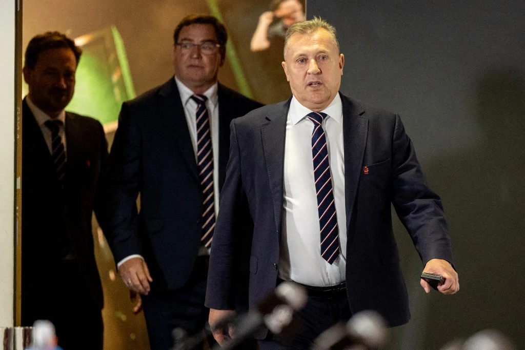 Cezary Kulesza po meczach z Holandią i Finlandią Trzech mężczyzn w garniturach i krawatach wychodzących z ciemnego pomieszczenia, na pierwszym planie mężczyzna o jasnych włosach trzymający przedmiot w ręku, w tle mikrofony sugerujące konferencję prasową.