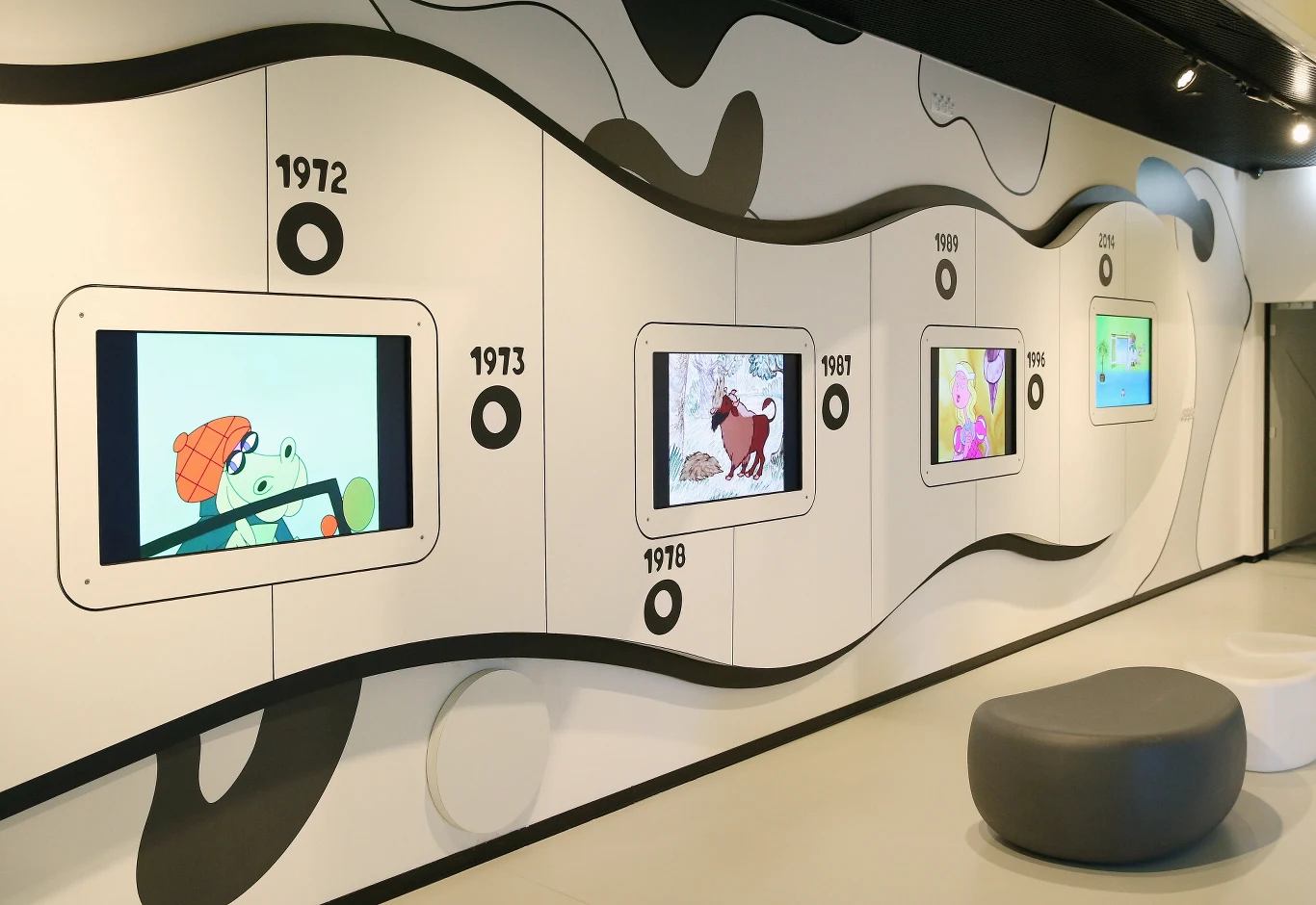 Multimedialna ekspozycja Studia Filmów Rysunkowych Ściana ilustrowana falistym wzorem z datami i ekranami wyświetlającymi kadry z animacji z różnych lat, obok znajduje się nowoczesne szare siedzisko.